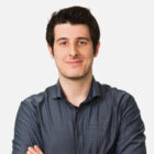 A portrait of a smiling man with short dark hair, wearing a dark blue shirt with a dotted pattern, against a white background.