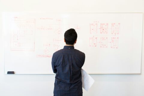 A person in a plaid shirt standing in front of a whiteboard with various red marker diagrams and flowcharts, holding a paper in their left hand.