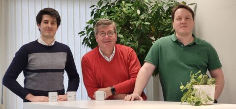 Drie mannen poseren bij een bureau met planten en koffiekopjes in een professionele kantooromgeving.