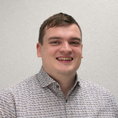 Man met een glimlach, gekleed in een kleurrijke blouse, staand tegen een lichte achtergrond.